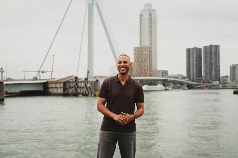 Luciano Currie lachend aan het water in Rotterdam, met de Erasmusbrug en hoge torens op de achtergrond.