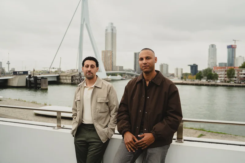 Seleman Arefi en Luciano Currie naast elkaar aan het water in Rotterdam, met de Erasmusbrug op de achtergrond.