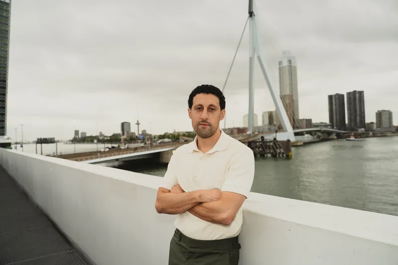 Seleman Arefi met de armen over elkaar aan een witte balustrade langs de Maas in Rotterdam, met de Erasmusbrug op de achtergrond.